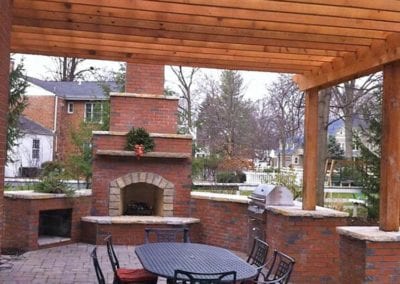 wood-pergola-covering-brick-patio-with-fireplace-in-background